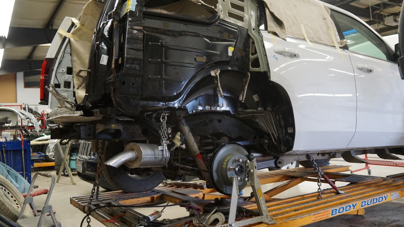 Rear view of a vehicle on a hoist inside a body shop with partial body removed. Many parts revealed, wheel, muffler, shocks and springs, etc.