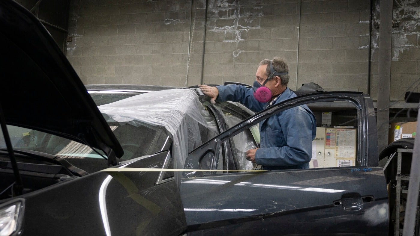 Man prepping a car for auto body repair. Wearing a face mask and utility overhauls is tapping plastic sheets to car inside auto body repair shop