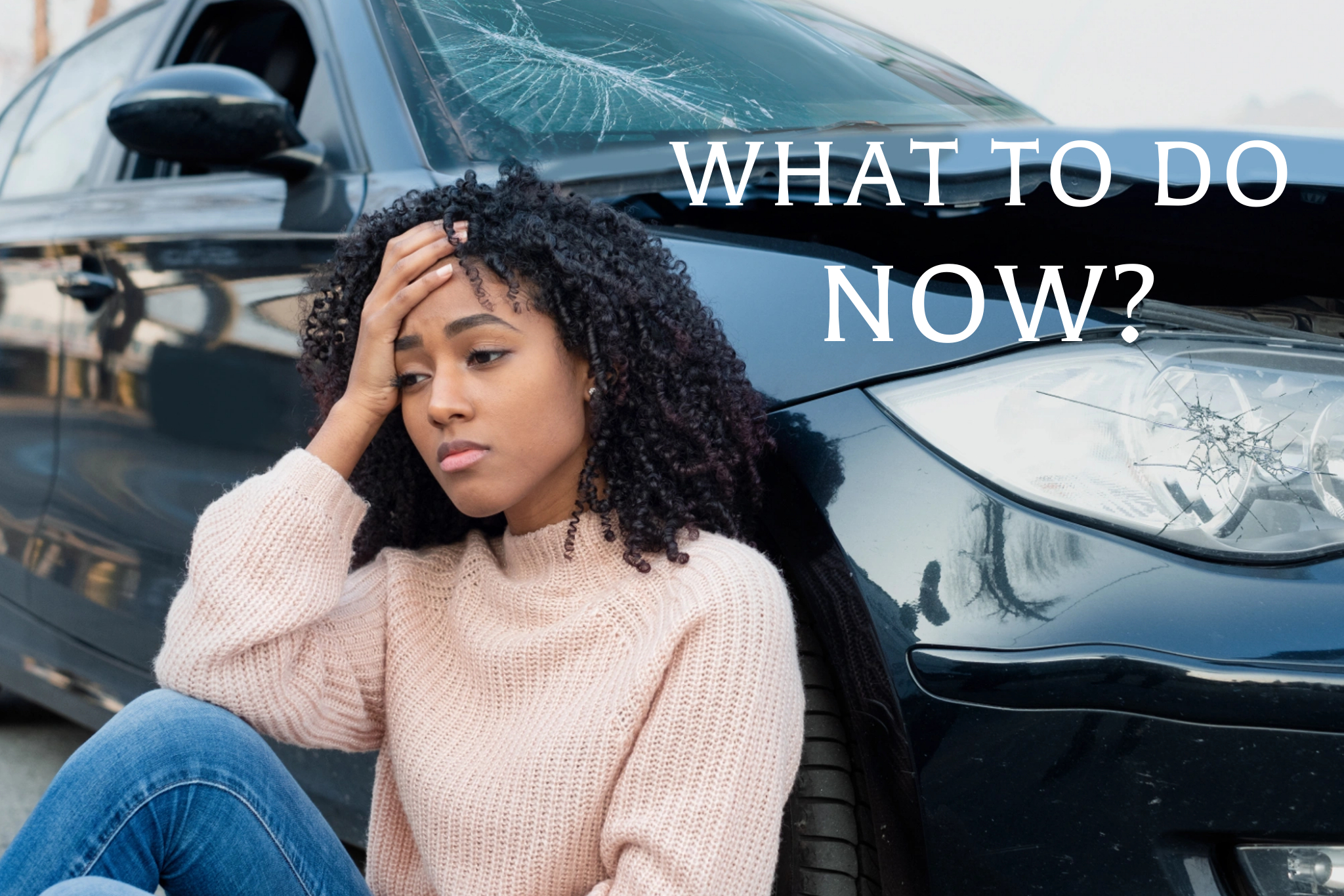 Young Woman sitting on ground lening her back against a damaged car with broken headlight and cracked widshield.